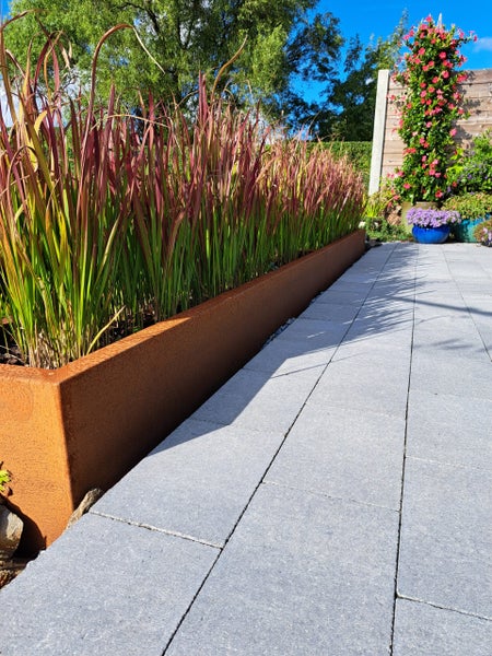 Hochbeet aus Cortenstahl mit rotem Ziergras neben einem Gartenweg aus grauen Terrassenplatten.