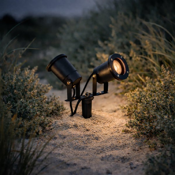 Zweiflammiger Gartenstrahler mit Erdspieß im Außeneinsatz in sandigem Gelände mit niedriger Bepflanzung.