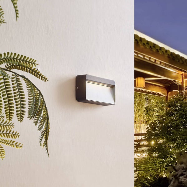 Moderne rechteckige LED-Außenwandleuchte in Anthrazit an einer Wand montiert, eingeschaltet, mit Garten im Hintergrund.