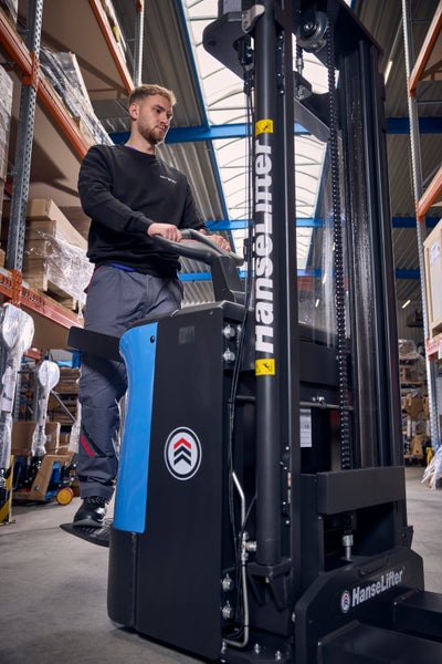 Ein Mann bedient einen elektrischen Hochhubwagen von HanseLifter in einem hellen, organisierten Lager mit hohen Regalen.