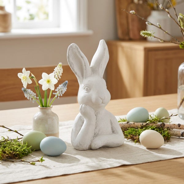 Dekorative Osterhasenfigur auf einem Holztisch mit Ostereiern, Moos und Frühlingsblumen in einer kleinen Vase.