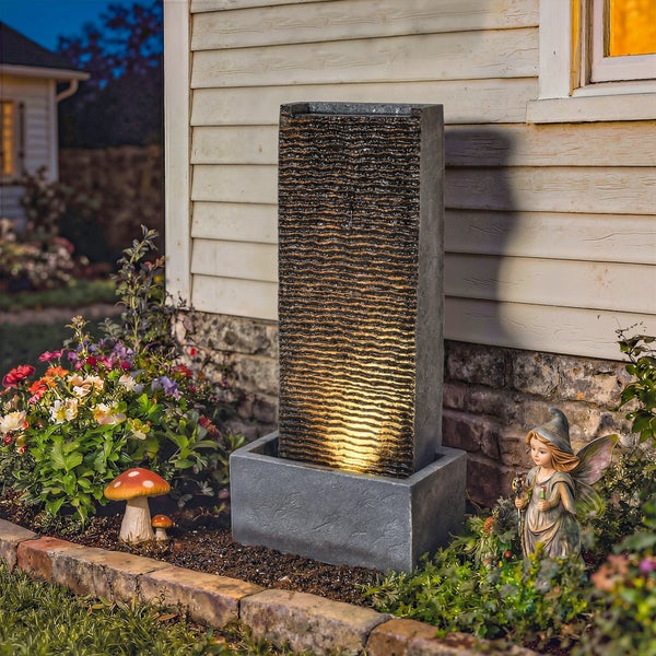 Rechteckiger Gartenbrunnen mit strukturierter Wellenoberfläche und LED-Beleuchtung in einem bepflanzten Gartenbeet.