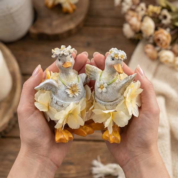 Zwei dekorative Enten-Figuren mit Blumenkränzen und gelben Stoffblumen in den Händen gehalten.