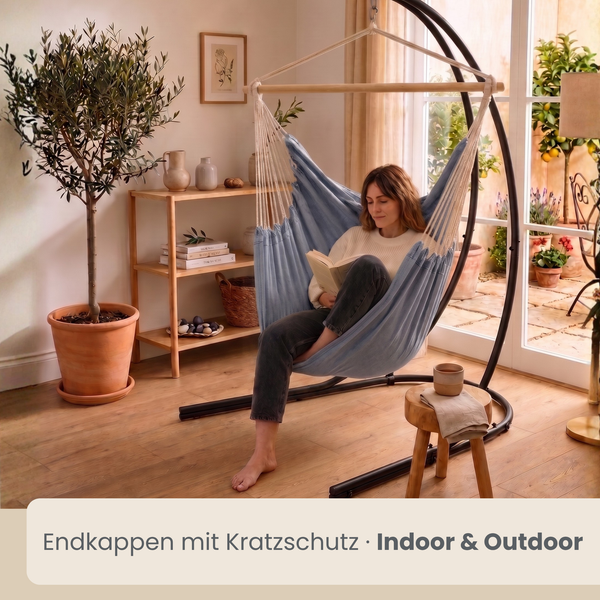 Eine Person sitzt in einem Hängesessel mit Gestell in einem wohnlichen Raum mit Olivenbaum und Holzboden. Ideal für den Innen und Außenbereich.