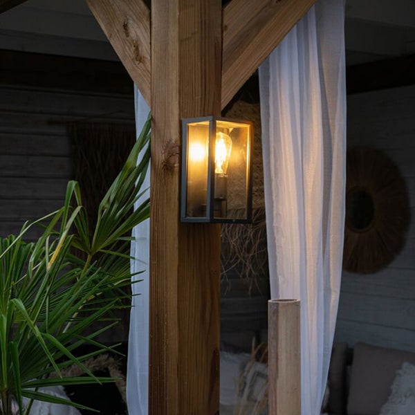 Schwarze Außenwandleuchte aus Metall und Glas im Laternen-Stil, montiert an einem Holzpfosten einer Terrasse, mit eingeschaltetem Leuchtmittel.