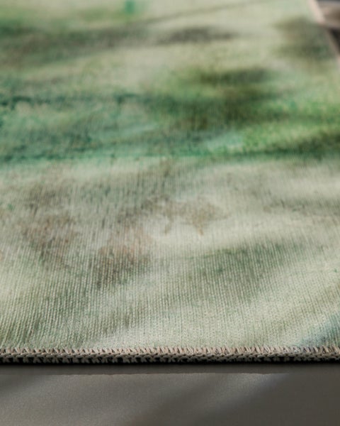 Detailansicht eines Teppichs mit abstraktem Muster in Grün und Beige sowie geketteltem Rand.