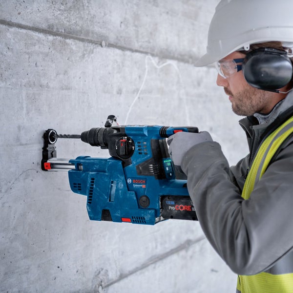 Ein Bauarbeiter bohrt mit einem blauen Bohrhammer ein Loch in eine Betonwand. Er trägt Schutzkleidung. Bosch Logo.