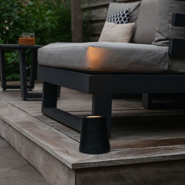Schwarze Außenleuchte auf einer Holzterrasse neben einem Loungesofa mit grauen Polstern bei Dämmerung.