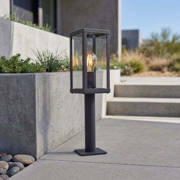 Moderne Sockelleuchte aus Metall in Anthrazit mit Glaslaterne und Filament-Leuchtmittel auf einem Betonweg neben einem Pflanzkübel.