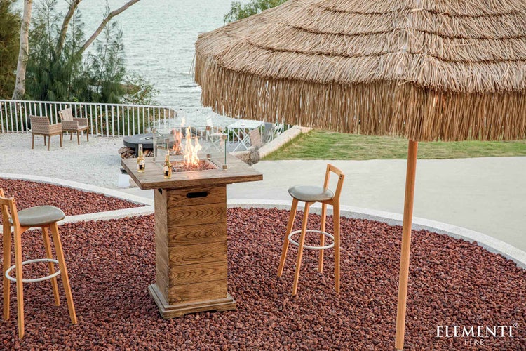 Outdoor-Stehtisch mit integrierter Feuerstelle in Holzoptik, Glasschutz und Barhockern auf einer Terrasse am Wasser.