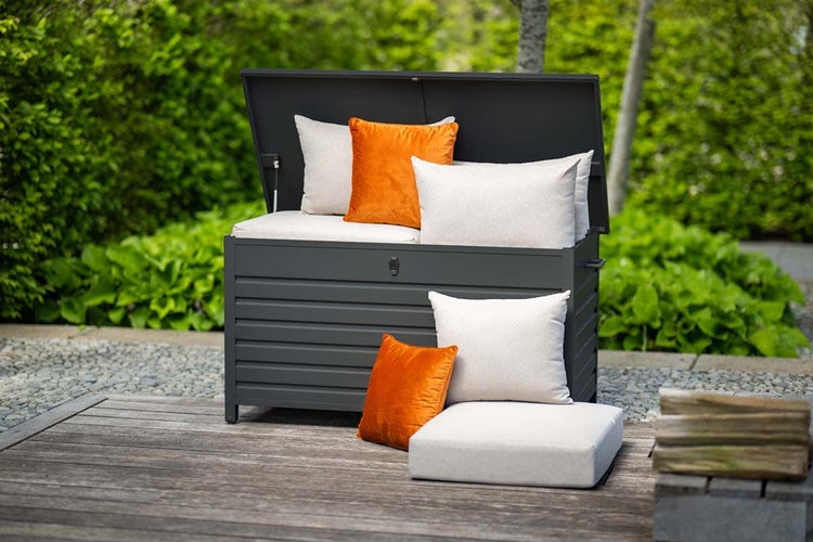 Dunkelgraue Kissenbox aus Metall mit offenem Deckel, gefüllt mit hellen und orangefarbenen Kissen auf einer Holzterrasse im Garten.