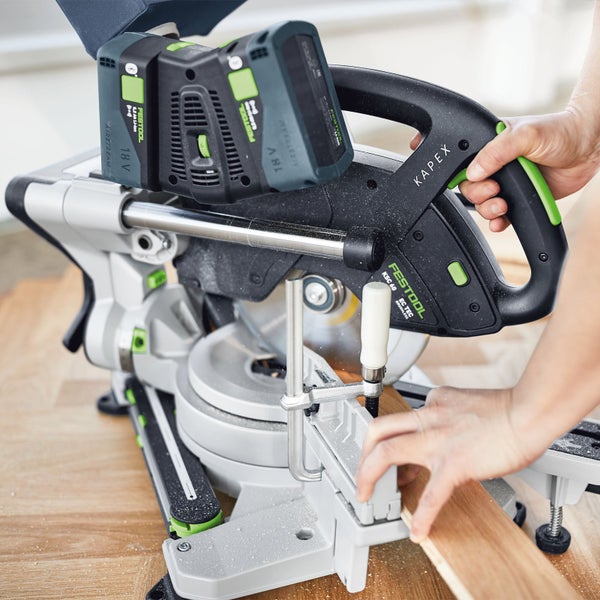 Eine Person nutzt eine Festool Kapex Akku-Kapp-Zugsäge mit zwei 18 Volt Akkus zum Sägen eines Holzbretts in einem hellen Arbeitsbereich.