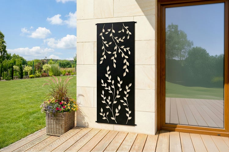 Dekoratives schwarzes Metallpaneel mit Blattmuster, montiert an einer hellen Steinwand auf einer Holzterrasse neben einem Pflanzgefäß im Garten.