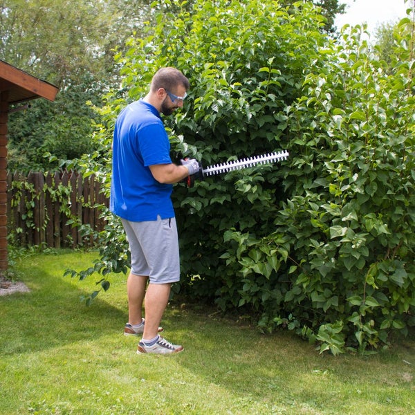 Mann mit Schutzausrüstung schneidet eine dichte grüne Hecke im Garten mit einer Heckenschere.