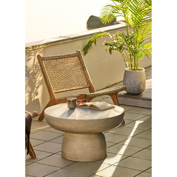 Runder Loungetisch in Betonoptik auf einer Terrasse neben einem Loungesessel aus Holz und Geflecht sowie einer Palme im Pflanzkübel.