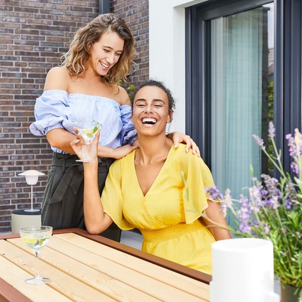 Zwei lachende Frauen auf einer Terrasse an einem Holztisch vor einer Ziegelwand und einem modernen Fensterrahmen.