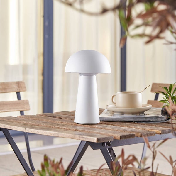 Weiße Tischleuchte in Pilzform auf einem hölzernen Klapptisch im hellen Außenbereich mit Kaffeetasse und Zeitschrift.