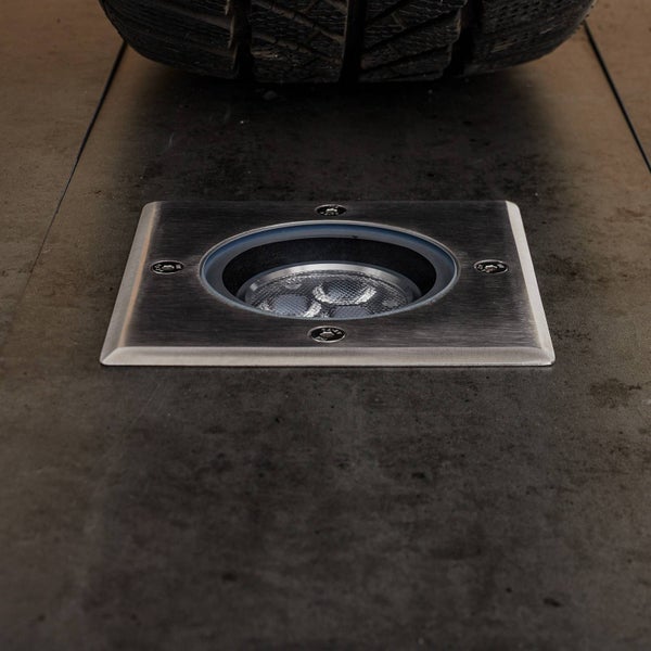 Quadratischer Bodeneinbaustrahler aus gebürstetem Edelstahl, befahrbar, montiert in einer befestigten Fläche unter einem Autoreifen.