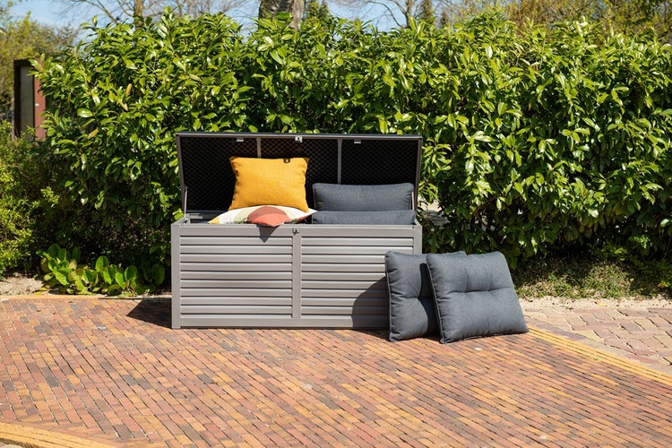 Offene Gartenbox aus Kunststoff mit Kissen auf einer gepflasterten Terrasse vor einer Hecke.
