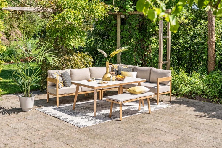 Gartenlounge-Set aus Holz auf einer Terrasse, bestehend aus Ecksofa, Esstisch und Sitzbank mit beigen Polstern inmitten von grünen Pflanzen.