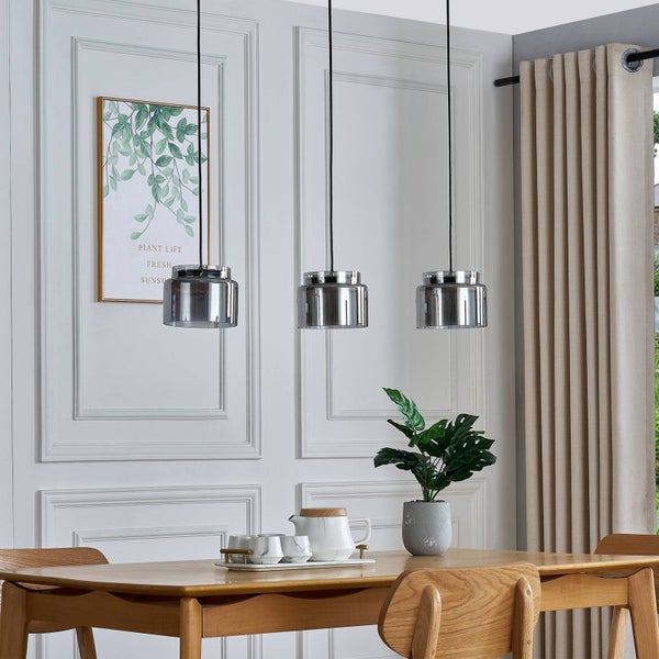 Drei moderne Pendelleuchten mit Glasschirmen über einem Esstisch aus Holz in einem hellen Raum mit Wandpaneelen und Zimmerpflanze.
