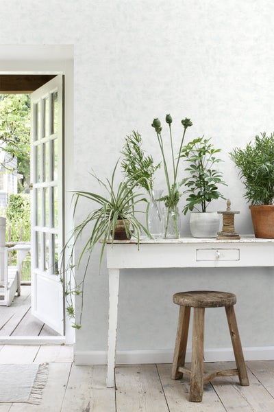 Helle Innenraumszene mit einem weißen Tisch, Pflanzen und einem hölzernen Hocker vor einer strukturierten Wand.