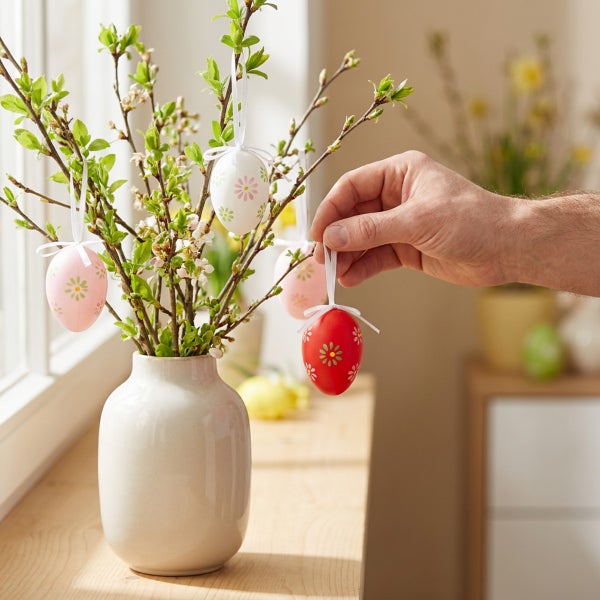 Eine Hand hängt ein österlich geschmücktes Osterei mit Blumenmuster an einen Zweig in einer Vase auf der Fensterbank.