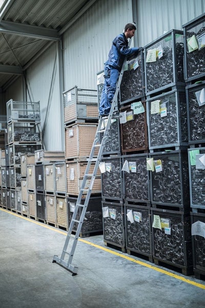 Mitarbeiter nutzt eine Aluminiumleiter in einem Lagerhaus mit hohen Gitterboxen für die Kommissionierung von Waren.