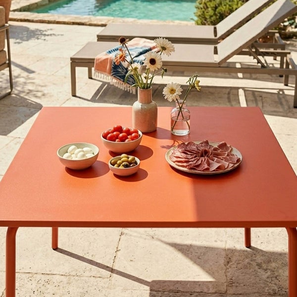 Moderner quadratischer Metall-Gartentisch auf einer hellen Terrasse am Pool, gedeckt mit Snacks und Blumen, Sonnenliegen im Hintergrund.