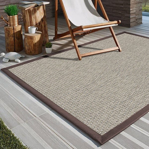 In- und Outdoor Teppich in Sisal-Optik auf einer hellen Terrasse mit Holz-Liegestuhl und rustikalen Beistelltischen aus Baumstämmen.
