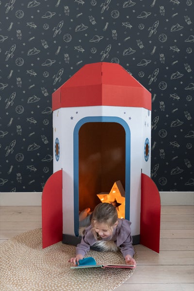 Kind liest in einem Raketen-Spielhaus auf einem Teppich vor einer Tapete mit Weltraummotiven