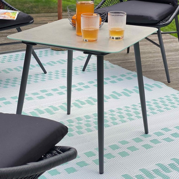 Moderner quadratischer Gartenbeistelltisch mit hellgrauer Platte und dunklen Metallbeinen auf einem gemusterten Outdoor-Teppich auf einer Terrasse.