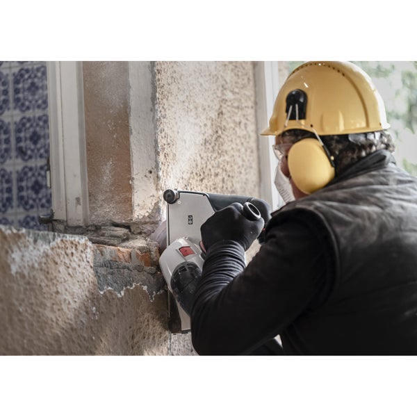 Ein Bauarbeiter verwendet eine Mauernutfräse an einer Wand, trägt dabei Schutzkleidung wie Helm, Gehörschutz, Schutzbrille und Maske.