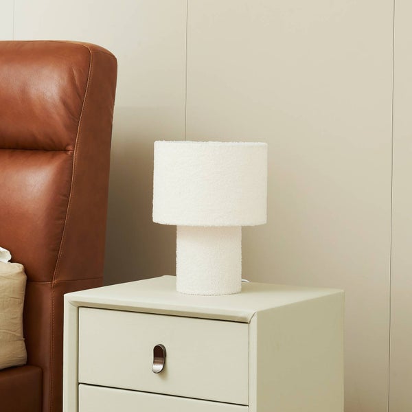 Weiße Tischleuchte mit Teddy-Stoff-Struktur auf einem hellen Nachttisch neben einem braunen Lederkopfteil eines Bettes.