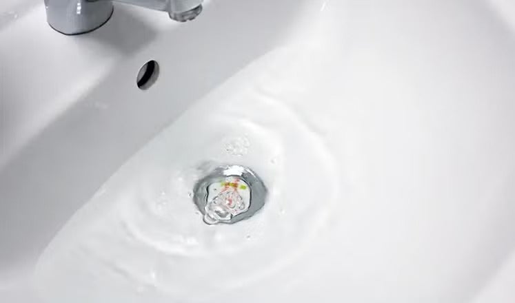 Ein weißes Waschbecken mit einem Metallabfluss, aus dem Wasser fließt, mit einem Teil eines Wasserhahns.