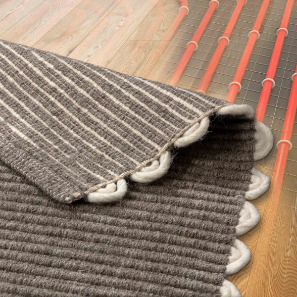 Gewebter Teppich auf Holzboden, teilweise umgeklappt, um darunterliegende Fußbodenheizungsrohre auf einem Gitter zu zeigen.