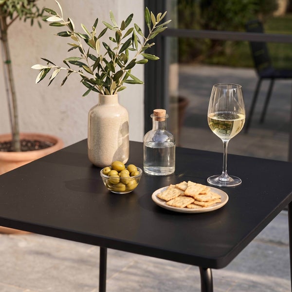 Quadratischer schwarzer Metalltisch auf einer hellen Terrasse mit einer Vase mit Olivenzweigen, einer Schale Oliven, Crackern und einem Glas Wein.