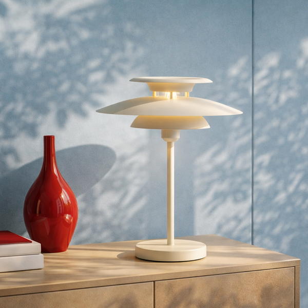 Moderne Tischleuchte mit mehrstufigem Schirm auf Holzkommode neben roter Vase vor blauer Wand mit Blätterschatten.