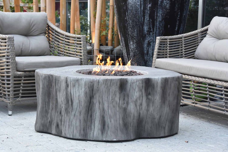 Gaskamin in Baumstamm-Optik mit Holzstruktur, offener Flamme und Lavasteinen zwischen zwei Loungesesseln auf einer Terrasse.