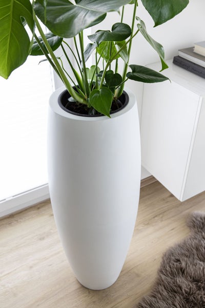 Hoher weißer Pflanzkübel mit einer Monstera-Pflanze in einem hellen, aufgeräumten Wohnraum auf Holzboden.