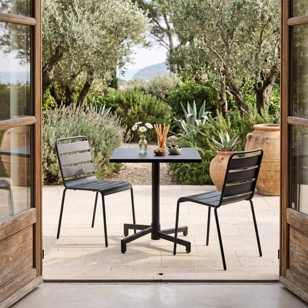 Modernes Bistro-Set aus schwarzem Metall auf einer hellen Steinterrasse, bestehend aus einem quadratischen Tisch und zwei Gartenstühlen.