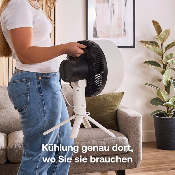 Eine Person hält einen modernen weißen Tischventilator mit Stativ in einem hellen, organisierten Wohnzimmer mit Stoffsofa und Zimmerpflanze.