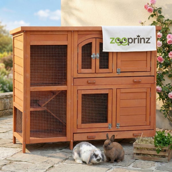 Großer zweistöckiger Kaninchenstall aus Holz mit mehreren Abteilen, Drahtgitter und Rampe im Außenbereich mit zwei Kaninchen und zooprinz Logo.