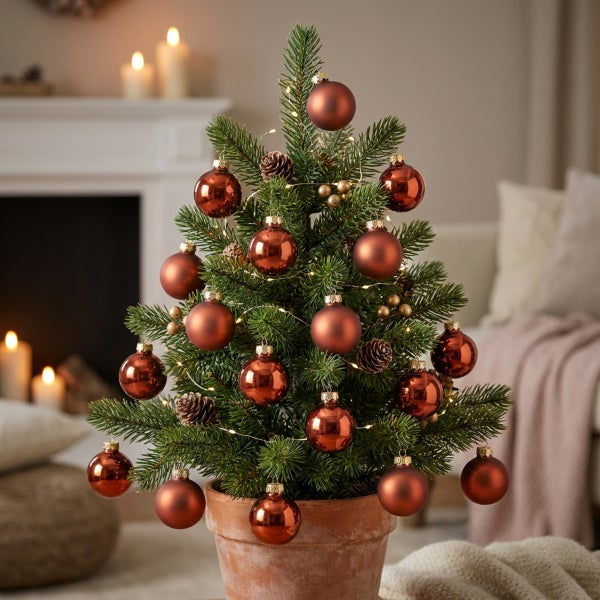 Ein kleiner Weihnachtsbaum im Tontopf geschmückt mit Weihnachtskugeln und Lichterkette vor einem Kamin in einer gemütlichen Wohnumgebung.