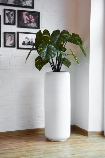 Hoher zylindrischer Pflanzkübel in Weiß mit Monstera-Pflanze in einem modern gestalteten Wohnbereich mit weißer Ziegelwand.