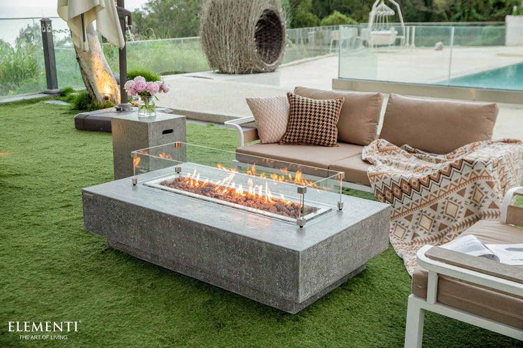 Moderner Gaskamin-Tisch in Betonoptik mit Glasschutz in einer Outdoor-Lounge mit Sofa und Pool. Elementi Logo.