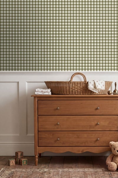 Kinderzimmer mit karierter Tapete, weißen Wandpaneelen, Holzkommode, Weidenkorb und Holzspielzeug auf Dielenboden.