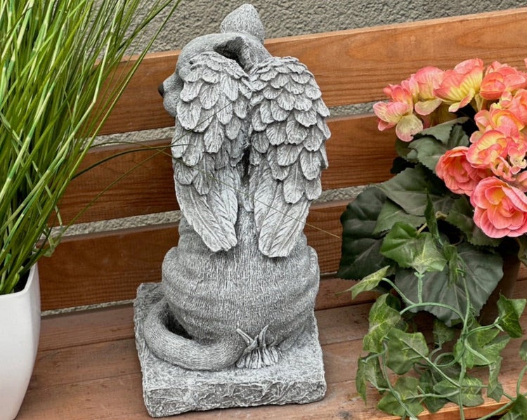 Tierengel-Gartenfigur mit Flügeln in Steinoptik von hinten auf einer Holzbank neben Pflanzen.