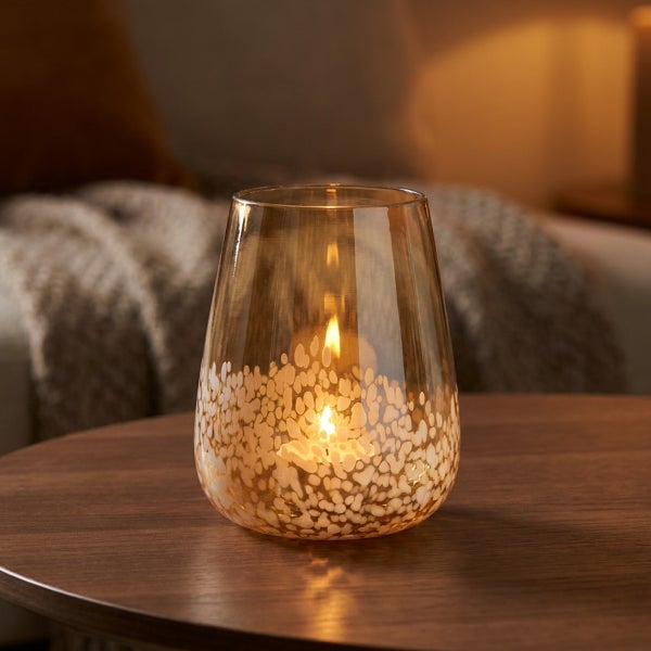 Ein gläsernes Windlicht mit Pünktchenmuster auf einem Holztisch in einem gemütlichen Wohnzimmer.