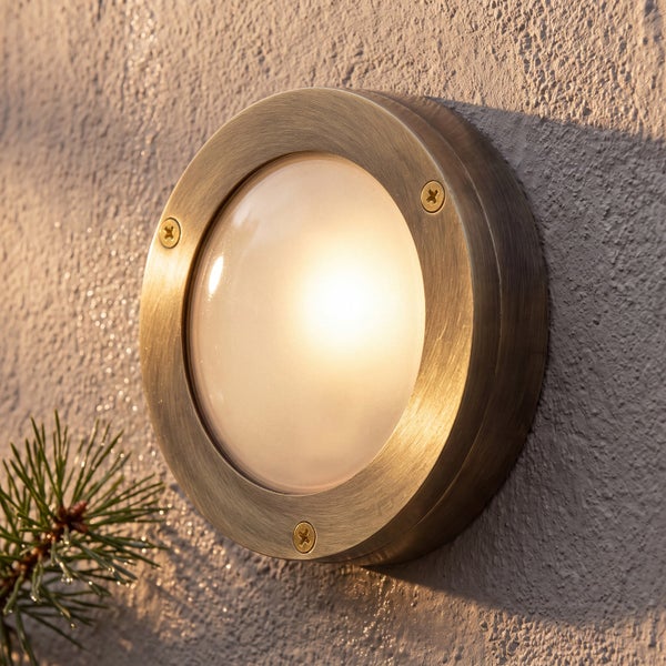 Runde Außenwandleuchte mit Rahmen aus gebürstetem Metall und mattiertem Glas, beleuchtet an einer strukturierten Fassade.
